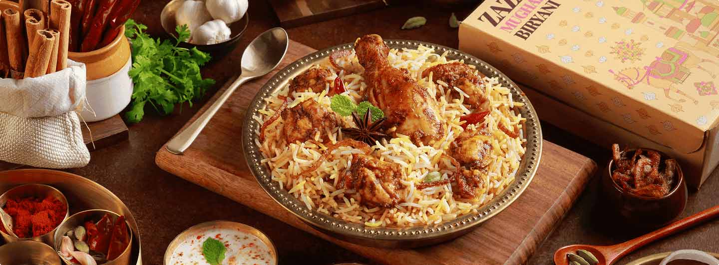 Freshly prepared ZAZA Mughal chicken biryani on a brass plate surrounded by aromatic whole spices, cinnamon, garlic, and chili peppers